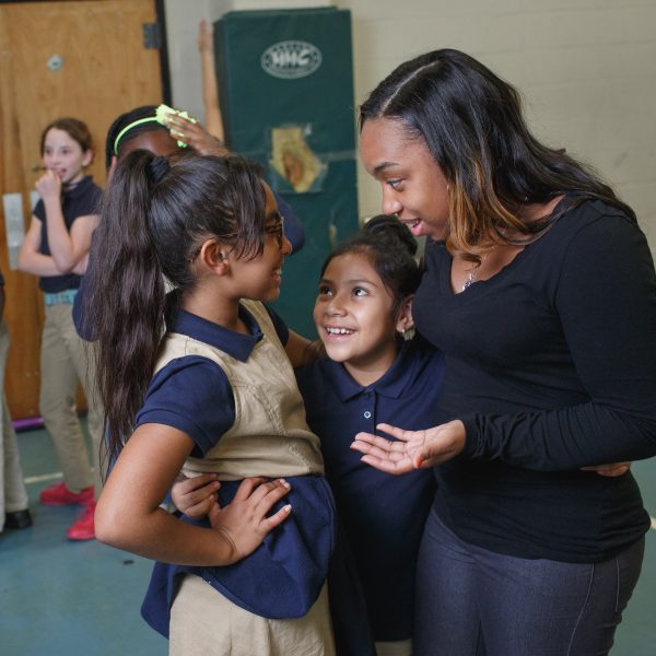 Two young female students look up to their teacher in a school gym, while their classmate line up behind them.