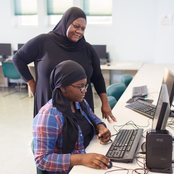 A loving mother stands over her young daughter, both concentrating on the computer screen.