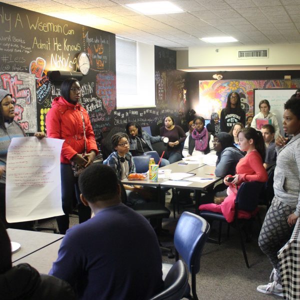 Proudly presenting their latest project, two teenagers stand before a group of their peers in a colorful youth center.