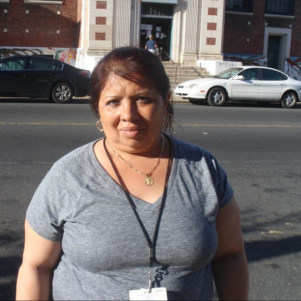 Smiling towards the camera, a middle-aged youth instructor stands proudly outside of a school in her neighborhood.