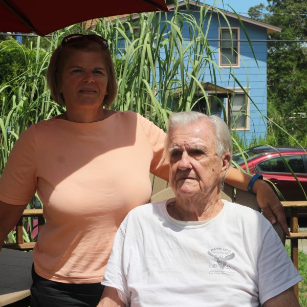 Proudly smiling towards the camera, an adult woman and her senior father enjoy summer weather on their porch.
