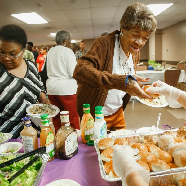 A young girl hands food to an elderly lady who is standing in line with friends in a busy dining hall.